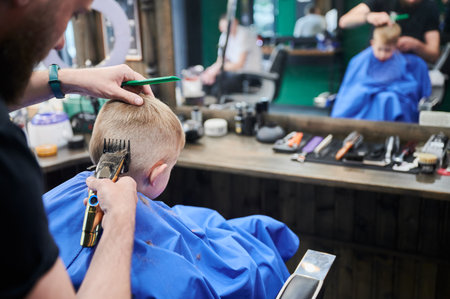 Barber using comb and shaver to cut hair. Back view of male hairdresser shaving little kids nape. Professional barber and cute boy in front of mirror.の写真素材