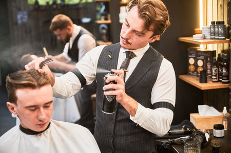 Professional barber using hairspray to finish hairstyle. Stylish hairdresser making final touches. Young customer getting fresh haircut.の写真素材