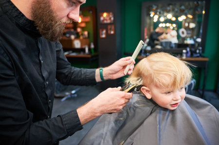 Professional hairdresser shaving boys head, using shaver and comb. Cute boy excited about new haircut. Blond boy getting stylish haircut in barbershop.の写真素材