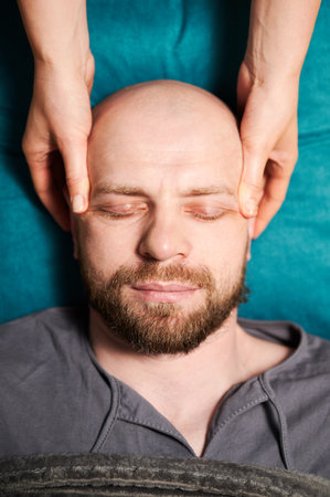 Close up of man getting thai face massage. Masseuse doing traditional massage treatment for male patient. Bald client with closed eyes, meditating, enjoying hand massage in calm atmosphere.の写真素材