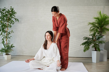 Masseuse making thai yoga massage. Woman therapist doing traditional massage treatment for female patient indoors. Client sitting on mat, meditating, enjoying hand massage in calm atmosphere.の写真素材