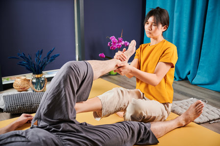 Masseuse making thai yoga massage. Woman therapist doing traditional massage treatment, stretching male patients body. Client lying on mat, enjoying hand massage in calm atmosphere.の写真素材