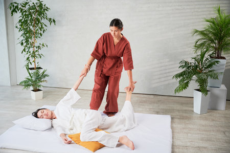 Masseuse making thai yoga massage. Woman therapist doing traditional massage treatment, stretching female patients body. Client lying on mat, enjoying hand massage in calm atmosphere.の写真素材