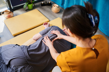 Masseuse making thai yoga massage. Woman therapist doing traditional massage treatment for male patient indoors. Client lying on mat, meditating, enjoying hand massage in calm atmosphere.の写真素材