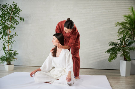 Masseuse making thai yoga massage. Woman therapist doing traditional massage treatment for female patient using elbow. Client sitting on mat, meditating, enjoying hand massage in calm atmosphere.の写真素材