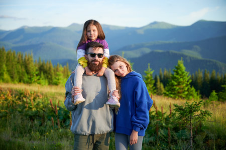 Family stands together in scenic meadow with backdrop of rolling mountains. Father carries his daughter on shoulders, both smiling and enjoying view, while mother stands beside them.の写真素材