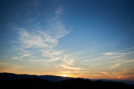 Sun sets behind rolling hills, casting warm, golden glow across sky. Wispy clouds, tinged with hues of orange and yellow, stretch across deepening blue sky. Serene and expansive landscape.の写真素材