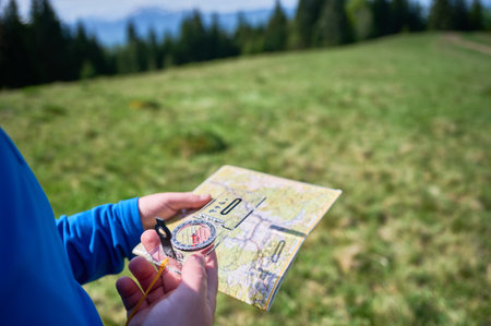 Person holds map and compass in green field. Scene features trees and distant mountains under partly cloudy sky. Focus on hands and navigation tools highlights act of orienting in nature.の写真素材