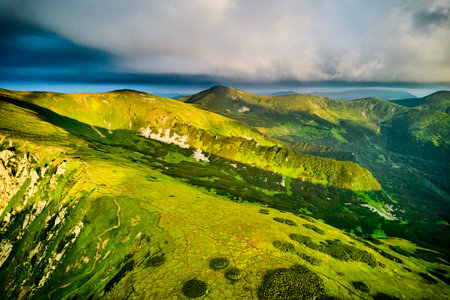 Breathtaking aerial view of green, rugged mountain peaks under dramatic sky. Scene captures contrast between vibrant landscape and moody clouds, with sunlight breaking through. Carpathians, Chornogoraの写真素材