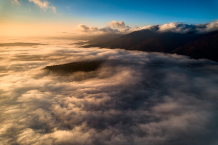 Aerial view of vibrant sunset over white dense clouds bathes in warm golden light, creating dreamlike landscape. Soft shadows play across undulating clouds, while distant landforms peek through mist.の写真素材