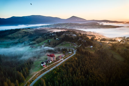 Aerial view of misty morning in mountains, with small village nestled among trees. Fog gently blankets landscape, creating serene and picturesque scene, as sun begins to rise in the background.の写真素材