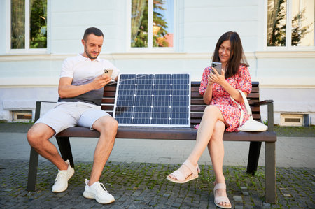 Man and woman sit on bench, smiling and using smartphones, connected to solar panel. Concept of sustainable renewable energy, demonstrating practical use of solar power for charging devices.の写真素材