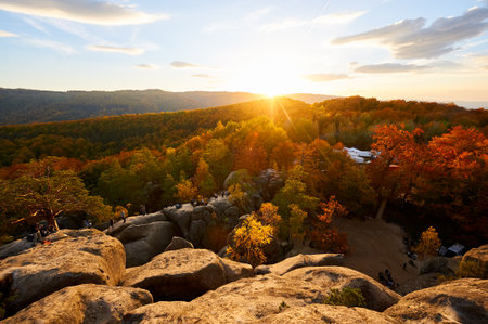 Setting sun casts golden glow over autumn landscape, illuminating vibrant red and orange foliage. Rugged rock formations in foreground under cloudy sky. Dovbush Rocks, Carpathian mountains, Ukraine.の写真素材