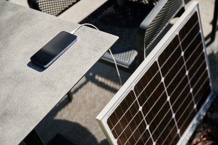 Smartphone connected to solar panel on outdoor table, emphasizing practical use of solar energy for charging devices. Integration of renewable energy, showcasing portable solar technology.の写真素材