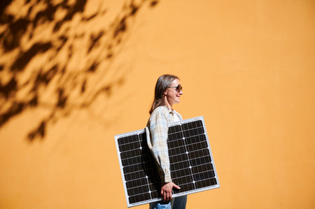 Woman holding solar panel for charging, stands against vibrant orange wall. Smiling girl wears sunglasses. Artistic shadows cast by nearby trees branches on wall.の写真素材