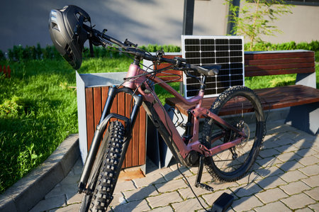 Pink electric mountain bike parked next to wooden bench, connected to solar panel for charging. Bike, with helmet hanging on handlebars, rests on paved area surrounded by greenery and grass.の写真素材