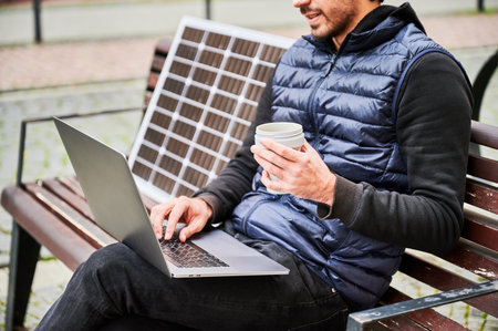 Cropped view of man sits on wooden bench in urban setting, working on laptop with cup of coffee in hand. Solar panel rests beside, emphasizing sustainable, mobile workspace.の写真素材