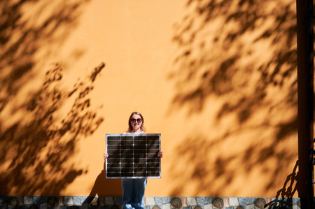 Woman holding solar panel for charging, stands against vibrant orange wall. Smiling girl wears sunglasses, white top, blue jeans. Artistic shadows cast by nearby trees branches on wall.の写真素材