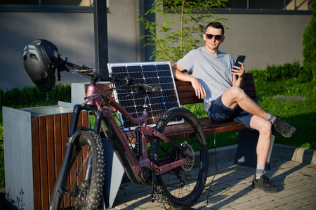 Man cyclist using solar panel for charging electric mountain bike outdoors. Guy in sunglasses sits on bench, holding phone, casually dressed in t-shirt, shorts, enjoying sunny outdoor setting.の写真素材