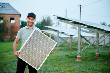 Man holds solar panel while standing in solar farm. Happy guy wearing black cap and light green polo...