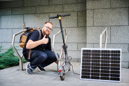 Man connects solar panel to electric scooter for charge, giving thumbs up. Integration of solar power as sustainable energy source for charging electric scooters, promoting eco-friendly transportationの写真素材