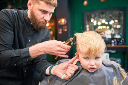 Professional hairdresser shaving boys head, using shaver and comb. Cute boy excited about new haircut. Blond boy getting stylish haircut in barbershop.の写真素材