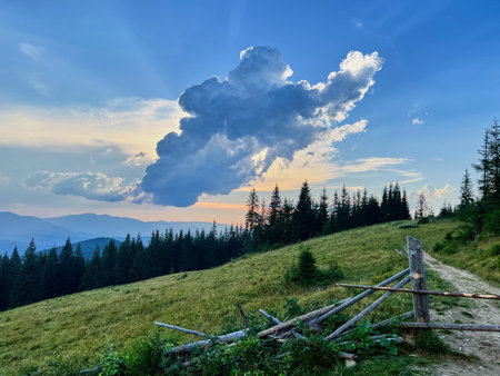 Dramatic cloud formation dominates evening sky over peaceful mountain landscape. Tall pine trees and grassy hill are in foreground, with rustic wooden fence and path leading into scene.の写真素材