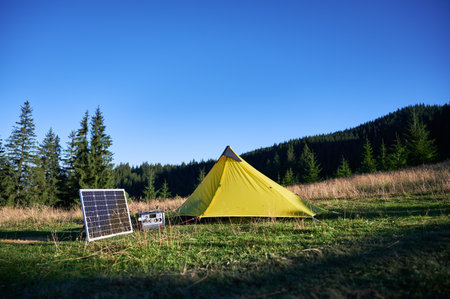Tourist tent with solar panel and portable power station nearby in grassy field. Scene surrounded by rolling hills, tall pine trees under clear blue sky, showcasing eco-friendly camping setup.の写真素材
