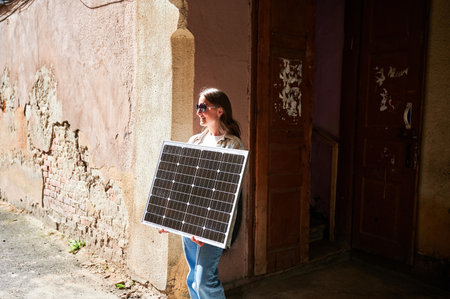Woman holding solar panel for charging, stands against weathered, textured wall. Smiling female in...