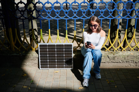 Happy woman using smartphone next to photovoltaic solar panel. Integration of sustainable renewable energy into everyday life, demonstrating practical use of solar power for charging devices.の写真素材