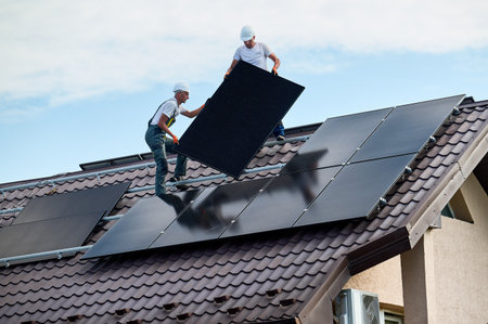 Workers building solar panel system on roof of house. Two men installers in helmets carrying...