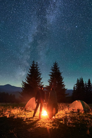 Two hikers stand by campfire under vast, star-filled sky, near tents glowing softly in background....