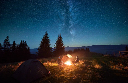 Silhouette of couple hikers resting by campfire under star-filled sky, near illuminated tents at campsite. Night camping in mountains, night sky create magical and peaceful atmosphere.の写真素材