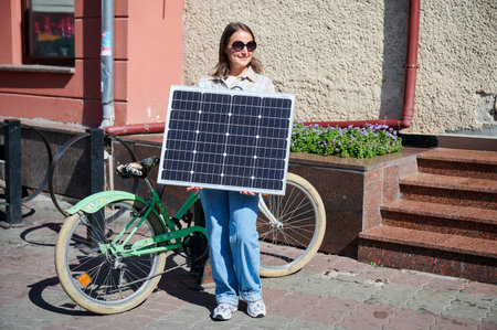 Smiling woman holding solar panel, standing beside green bicycle. Happy female in sunglasses and...