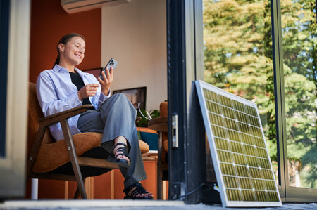 Happy woman using smartphone connected to photovoltaic solar panel. Integration of sustainable renewable energy into everyday life, demonstrating practical use of solar power for charging devices.の写真素材