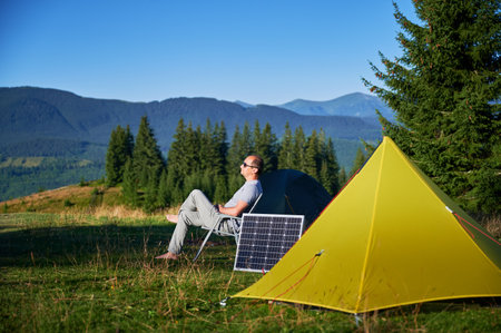 Man relaxes in folding chair, soaking in sun beside solar panel and yellow tent. Green trees and majestic mountains under clear blue sky, highlighting an eco-friendly camping experience.の写真素材