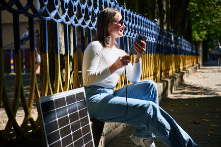 Happy woman connecting smartphone to photovoltaic solar panel. Integration of sustainable renewable energy into everyday life, demonstrating practical use of solar power for charging devices.の写真素材