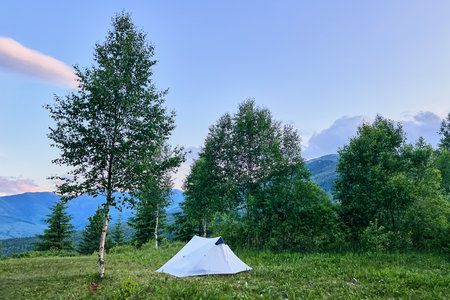 White tourist tent on grassy clearing, surrounded by lush trees with distant blue mountains under sky tinged with pink clouds. Peaceful scene exudes tranquility of nature and serene mountain camping.の写真素材