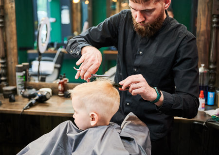 Man barber cutting little boys hair using comb and scissors. Child getting haircut from adult male, likely barber. Professional hairdresser and cute client at modern barbershop.の写真素材