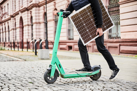 Cropped view of man rides electric scooter while carrying solar panel. Integration of solar power as sustainable energy source for charging electric scooters, promoting eco-friendly transportation.の写真素材