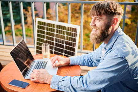 Man works on laptop and smartphone at outdoor table, with glass of iced coffee. Solar panel nearby, emphasizing sustainable, modern eco-friendly workspace that combines technology and sustainability.の写真素材