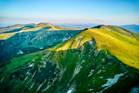 Aerial view of rugged mountain range with lush green slopes at sunset. Landscape showcases beauty of untouched nature with distant peaks and valleys bathed in soft sunlight.の写真素材