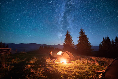 Hiker woman relaxes by campfire, illuminated by its warm glow, with tent nearby under stunning, star-filled sky. Milky Way stretches overhead, adding magical touch to serene mountain setting.の写真素材