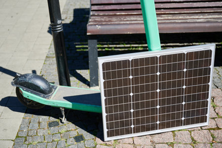 Mint-green electric scooter rests on cobblestone path, with solar panel leaning against it. Solar...