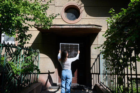 Woman holding photovoltaic solar panel in front of historical building, wearing plaid shirt and sunglasses. Concept of integration of sustainable renewable energy sources into old architecture.の写真素材