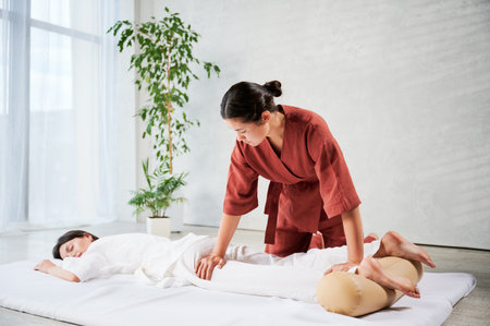 Masseuse making thai yoga massage. Woman therapist doing traditional massage treatment for female patient indoors. Client lying on mat, meditating, enjoying hand massage in calm atmosphere.の写真素材