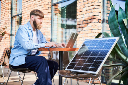 Man works on laptop at outdoor round table, with glass of iced coffee. Solar panel nearby, emphasizing sustainable, modern eco-friendly workspace that combines technology and sustainability.の写真素材
