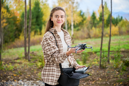 In a beautiful forest during autumn, a smiling young woman stands proudly holding a drone. Colorful...