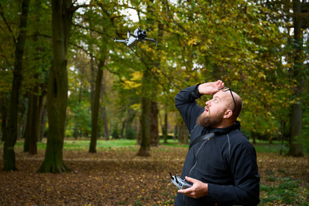 A man with a beard operates a drone in a lush park filled with colorful autumn leaves. He appears...