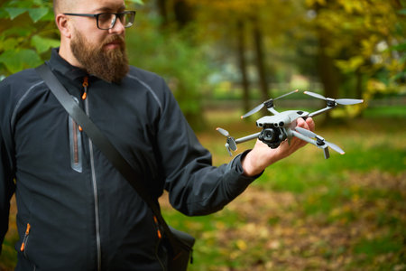 A man stands outdoors in a vibrant park, holding a drone in his hand. The trees around him display...